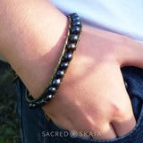 A person's forearm showcasing a black and blue pearl bracelet with bronze leather accents and an antique silver button.