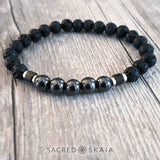 The I Am Courageous bracelet from Sacred Skaia is black, brown and silver and features hematite, bronzite and lava stone crystals with silver accents and is ideal for all genders, shown on a weathered wood background.